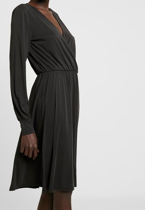 Kvinde iført en sort, knælange kjole med lange ærmer, V-hals og indsnøring i taljen, stående mod en ensfarvet lys baggrund.