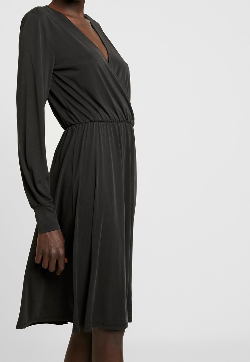 Femme portant une robe noire à manches longues, longueur genou, avec un col en V et une taille froncée, debout devant un fond clair uni.