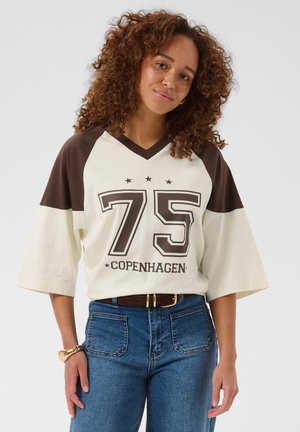 Femme aux longs cheveux bouclés portant une chemise crème et marron avec l’inscription « 75 Copenhagen », un jean bleu et un bracelet doré, debout devant un fond uni.