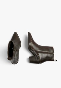 Botas de tobillo de cuero marrón oscuro con cremalleras laterales, tacones apilados, punteras puntiagudas y detalles de tachuelas de latón a lo largo de los bordes, exhibidas sobre un fondo blanco.