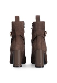 Brown suede ankle boots with a high heel, featuring a side zipper, decorative straps, and a logo accent on the heel base.
