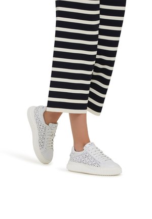Witte sneakers met een decoratief geperforeerd ontwerp, rubberen zool en gestructureerde details. Gestreepte wijde broeken zijn zichtbaar op de achtergrond.