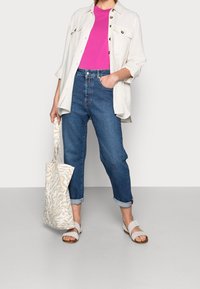 Camisa botón beige claro sobre una camiseta rosa brillante, combinada con jeans azules arremangados y sandalias blancas. Bolso de mano con cremallera de patrón de cebra.