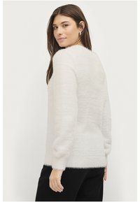 Weißer, flauschiger Pullover mit weicher Textur, langen Ärmeln und rundem Halsausschnitt, kombiniert mit schwarzen Hosen. Der Stoff hat einen leichten Glanz.