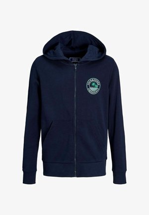 Personne portant un sweat à capuche zippé bleu marine avec un logo circulaire, un pantalon noir et des baskets noires et blanches, debout devant un fond gris uni.