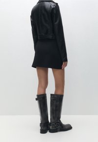 Černá kožená krátká bunda, černé přiléhavé šaty, kozačky ke kolenům se sponami, matný povrch, hladká textura, těsně přiléhající střih.