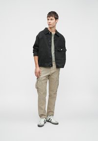 Ung mand står iført en sort denimjakke, lysegrøn T-shirt, beige cargo-bukser og hvide sneakers med sorte striber.