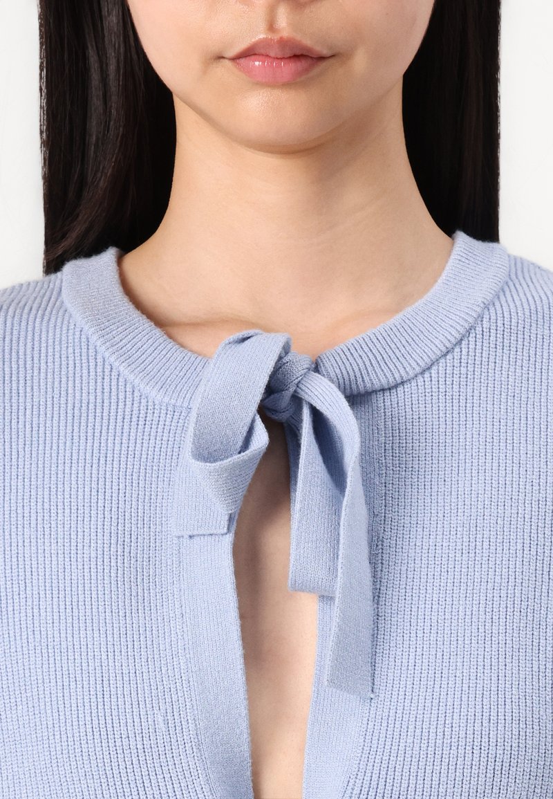 Jasnoniebieski sweter dzianinowy z okrągłym dekoltem, z wiązaniem w formie kokardy przy rozcięciu oraz prążkowaną fakturą na całej powierzchni.