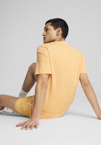 Maglietta corta a maniche corte arancione chiaro con collo a giro, abbinata a pantaloni corti gialli. Il modello è seduto e si volta leggermente all'indietro.
