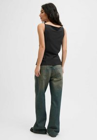 Kvinna med lång mörk hår som bär en svart ärmlös topp och löst sittande urblekta blå jeans, stående vänd bort från kameran mot en enfärgad vit bakgrund.