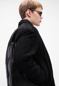 Giovane uomo con giacca in suede e pelle nera che indossa occhiali da sole, mostrato di lato su uno sfondo bianco semplice.