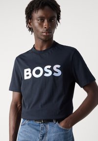Tamsiai mėlyna trumpomis rankovėmis medvilninis marškinėliai su dideliu, perėjimo sidabriniu "BOSS" logotipu ant krūtinės. Derinama su šviesiai mėlynomis džinsinėmis kelnėmis.