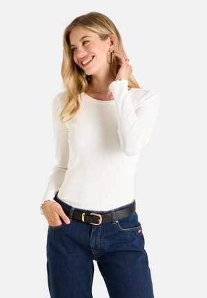 Lächelnde Frau trägt ein weißes langärmeliges Oberteil mit wellenförmigen Kanten, dunkelblaue Jeans, schwarzen Gürtel und Goldschmuck, posiert mit der Hand am Ohr.