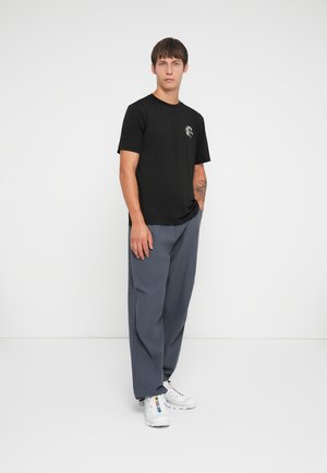 Must beebi lühikeste varrukatega must puuvillane T-särk, millel on graafiline disain rinnal, kombineeritud hallide vabade pükste ja valgete tennistega.