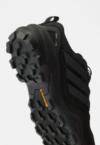 Čierna športová obuv s textúrovanou gumovou podrážkou s hlbokými výčnelkami pre trakciu, zosilnenou špičkou, sieťovinovým zvrškom a výraznými značkovými detailmi.