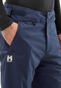 Pantalons de plein air bleu marine en matériau résistant à l'eau, avec une ceinture en Velcro, des poches latérales et un logo de marque visible.