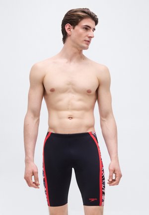 Paita päällään mustat ja punaiset polvenmittaiset uimahousut käyttävä mies, joka seisoo rennosti käsivarret sivuilla ja katsoo sivulle yksiväristä taustaa vasten.