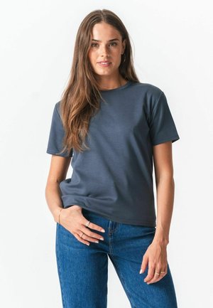 Kvinde med langt brunt hår iført en ensfarvet mørkegrå kortærmet t-shirt og blå jeans, stående med den ene hånd i lommen.