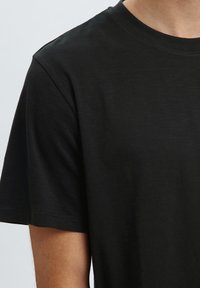 Primer plano de una persona que lleva una camiseta negra lisa de manga corta y cuello redondo sobre un fondo gris claro.