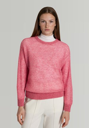 Joven mujer con el cabello liso y castaño, vistiendo un suéter de punto rosa sobre un cuello alto blanco y pantalones blancos, con un fondo gris liso.