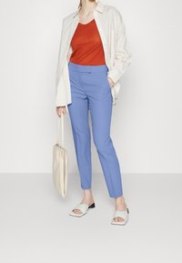 Ljust blå skräddarsydda byxor, orange tanktop och en oversized vit skjorta med knappar. Håller en krämfärgad handväska och bär ljusa sandaler.