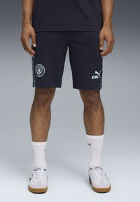 Mørkeblå sports shorts lavet af stof, med Manchester City logo, Puma branding og sidelister i lys blå. Hvide crew sokker og sneakers.