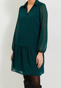 Teal dress with long sheer sleeves, V-neck collar, patterned texture, gathered waist, knee-length hem, paired with black thigh-high boots.