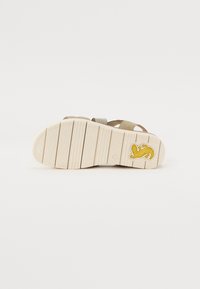 Sandaler med beige stoffremmer, en teksturert kremgummisåle og en lys gul logo-detalj på hælen. Har et flatt design og solid konstruksjon.