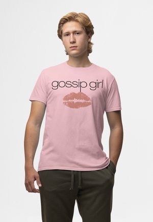 Jeune homme debout les mains dans les poches portant un t-shirt rose « gossip girl » avec une empreinte de lèvres sur fond blanc.