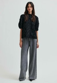 Blusa nera texture con ricami floreali, maniche a sbuffo corte e chiusura con bottone, abbinata a pantaloni in denim grigi a gamba larga e tacchi neri a punta.