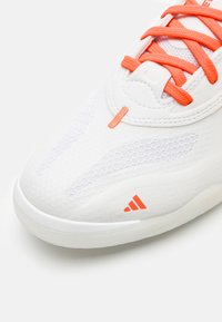 Zapatilla deportiva blanca con acentos naranjas, parte superior de malla texturizada y diseños suaves contrastantes. Presenta cordones y un detalle del logo en el lateral.