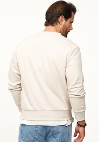 Hellbeige Sweatshirt mit Rundhalsausschnitt, gerippten Bündchen und Saum. Verfügt über eine glatte Textur und eine lockere Passform, getragen über einem weißen Shirt.