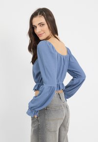 Top blu textured a maniche a palloncino con orlo arricciato, abbinato a jeans in denim grigio a vita alta, visto da dietro.