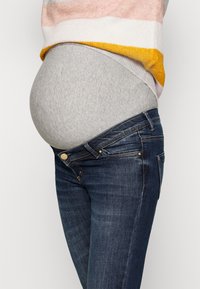 Graues T-Shirt unter einem mehrfarbig gestreiften Pullover, kombiniert mit unverschlossenen dunkelblauen Jeans, die einen auffälligen runden Bauch zur Schau stellen.