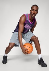 Košarkaš u ljubičastoj majici s natpisom "Vukovi", sivi šorts, drži narančastu košarkašku loptu, nosi crne tenisice i bijele čarape.