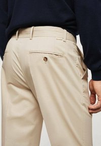 Pantaloni beige con una trama liscia, passanti per cintura e una tasca posteriore con bottone decorativo. Presentano una vestibilità sartoriale e tasche laterali.