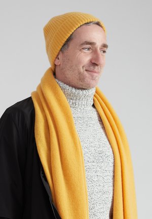 Geel geribbelde beanie en sjaal gecombineerd met een grijze gebreide coltrui. De sjaal hangt over het zwarte materiaal van de jas.