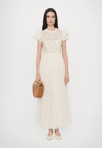 Crème-kleurige lange jurk met een met bloemen geborduurde korsage, pofmouwen en een geplooide rok. Model houdt een rieten handtas vast en draagt lichte schoenen.