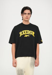 Melna īsām piedurknēm T-kreklu ar lielu dzeltenu "REEBOK" logo. Apaļš kakls, brīvs piegriezums, mīksta kokvilnas audums. Minimālistisks dizains, bez rakstiem.