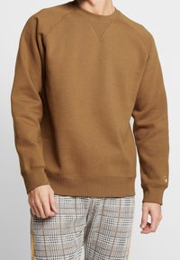 Brun sweatshirt med rund halsringning och ribbade muddarn, med en logotyp på ärmen, kombinerad med rutiga byxor.