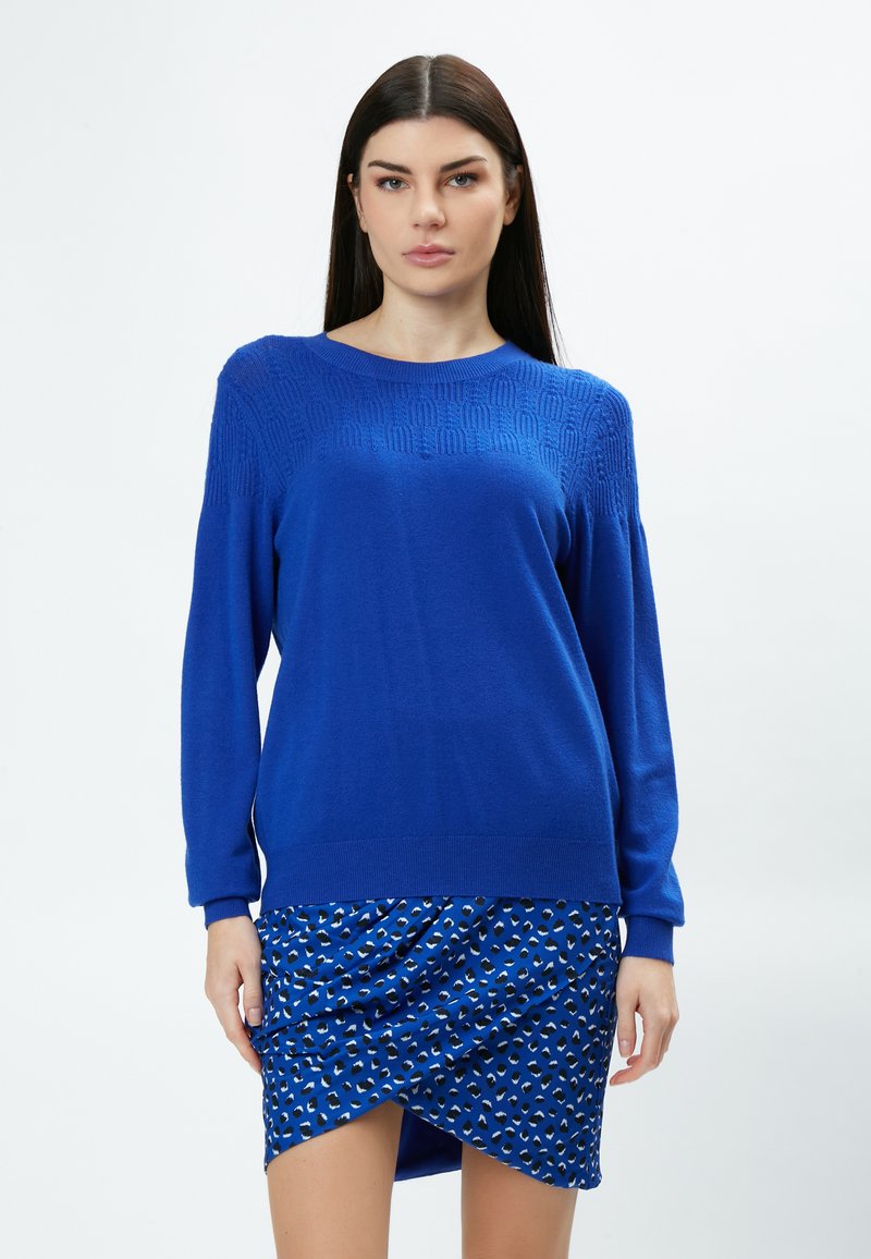 Blauer gestrickter Pullover mit einem strukturierten Muster an den Schultern, kombiniert mit einem blauen Rock, der ein kontrastierendes Muster aus kleinen Formen aufweist.