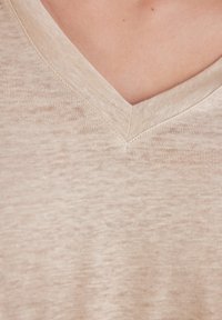 Beige top s V-výstřihem vyrobený z měkké, texturované tkaniny. Výstřih má čisté zakončení s jemnými variacemi v barvě a struktuře.