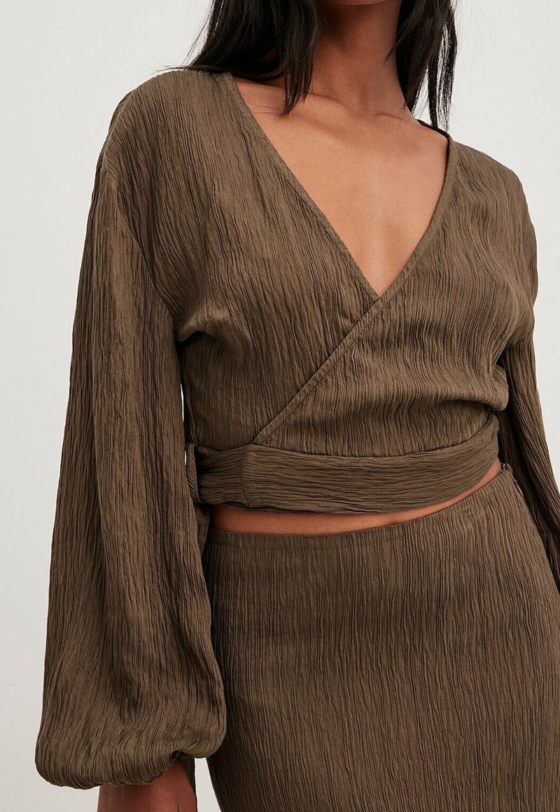 Femme portant un haut court cache-cœur en tissu marron texturé avec manches longues bouffantes et jupe taille haute assortie, sur un fond uni.