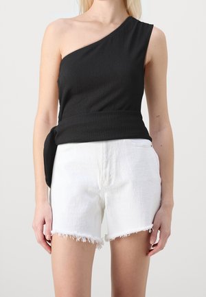 Frau trägt ein schwarzes ärmelloses Top mit einem Schulterriemen und weiße hoch taillierte Jeansshorts mit ausgefransten Säumen, stehend vor einem schlichten Hintergrund.