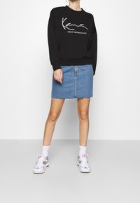 Persona con sudadera negra con texto blanco, falda de mezclilla azul con dobladillo deshilachado, calcetines blancos y zapatillas blancas de pie contra un fondo blanco.