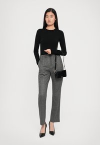 Schwarzes langärmeliges Oberteil kombiniert mit grauen Hahnentrittmuster-Hosen mit hoher Taille. Das Model hält eine kleine schwarze Handtasche und trägt schwarze spitze Absätze.