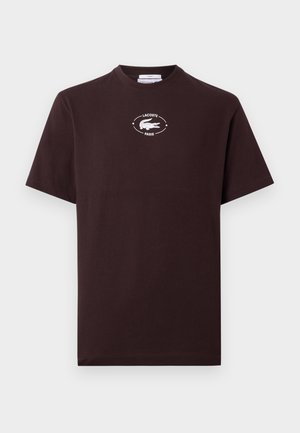 Un homme et une femme debout côte à côte, tous deux portant des t-shirts noirs à logo ; l'homme porte un pantalon blanc et des chaussures vertes, la femme une jupe plissée beige et des baskets noires.