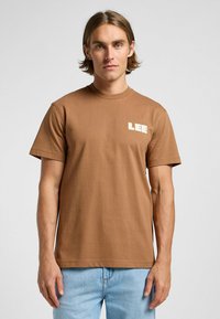 Brun bomulls t-shirt med rund hals och korta ärmar. På vänster bröst syns en kontrasterande vit "LEE"-logotyp.