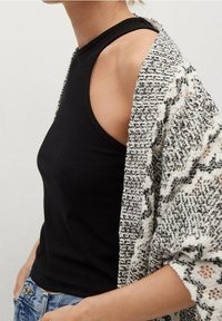 Donna che indossa un top senza maniche a coste nero, jeans azzurri chiari e un cardigan lavorato a maglia largo con un motivo bianco e nero.