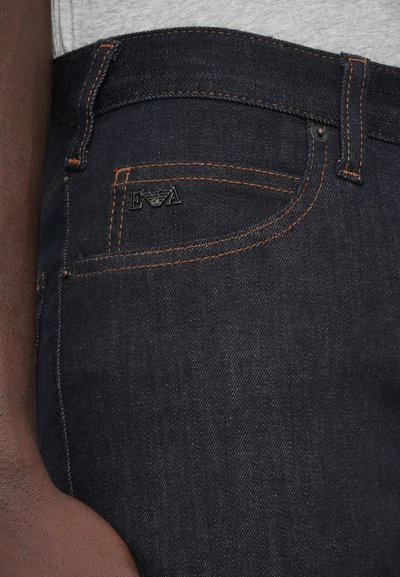 Tmavomodré riflové nohavice s oranžovým prešitím, s predným vreckom a čiernym detailom s logom. Hladká textúra a štandardný strih.